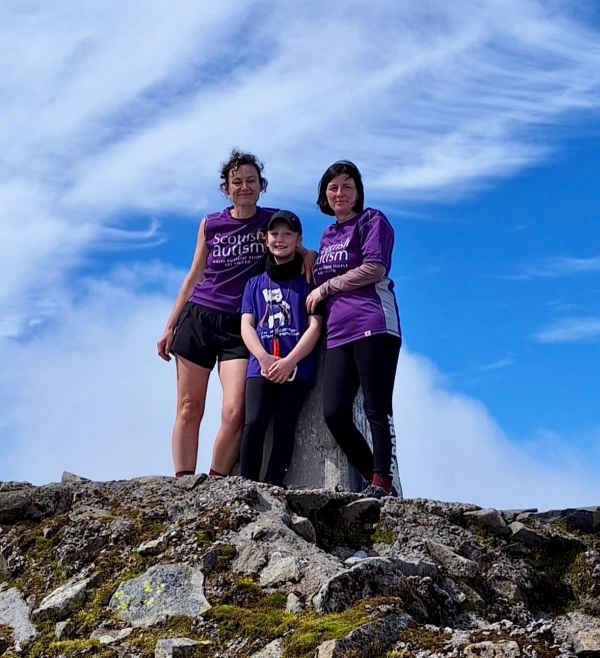 Image of two adults and a child, wearing Scottish Autism supporter tshirts, standing at the summit of a mountain with a blue sky background