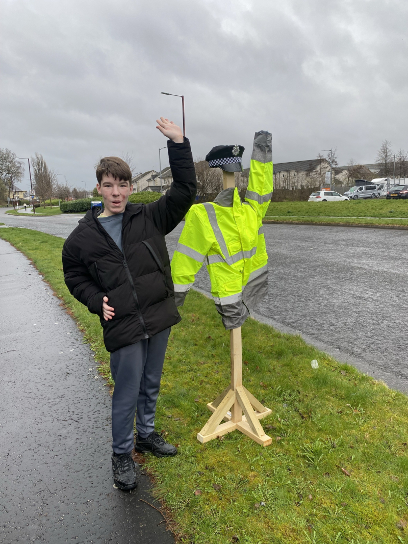 A photograph of Caine Arnott standing next to PC Struan a wooden structure with a police jacket and hat. Caine is holding up his arm