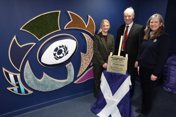 Image of three people smiling to camera as the Wooden Spoon Sensory Space is opened, the Scottish Rugby logo is on the wall, made from different tactile textured materials