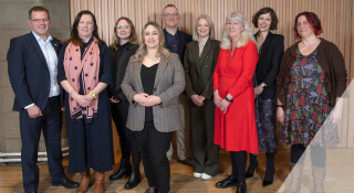 A group photo of nine individuals standing in front of a wooden paneled wall. They are all dressed in business or smart casual attire at Scottish Autism's Centre launch, smiling at the camera.